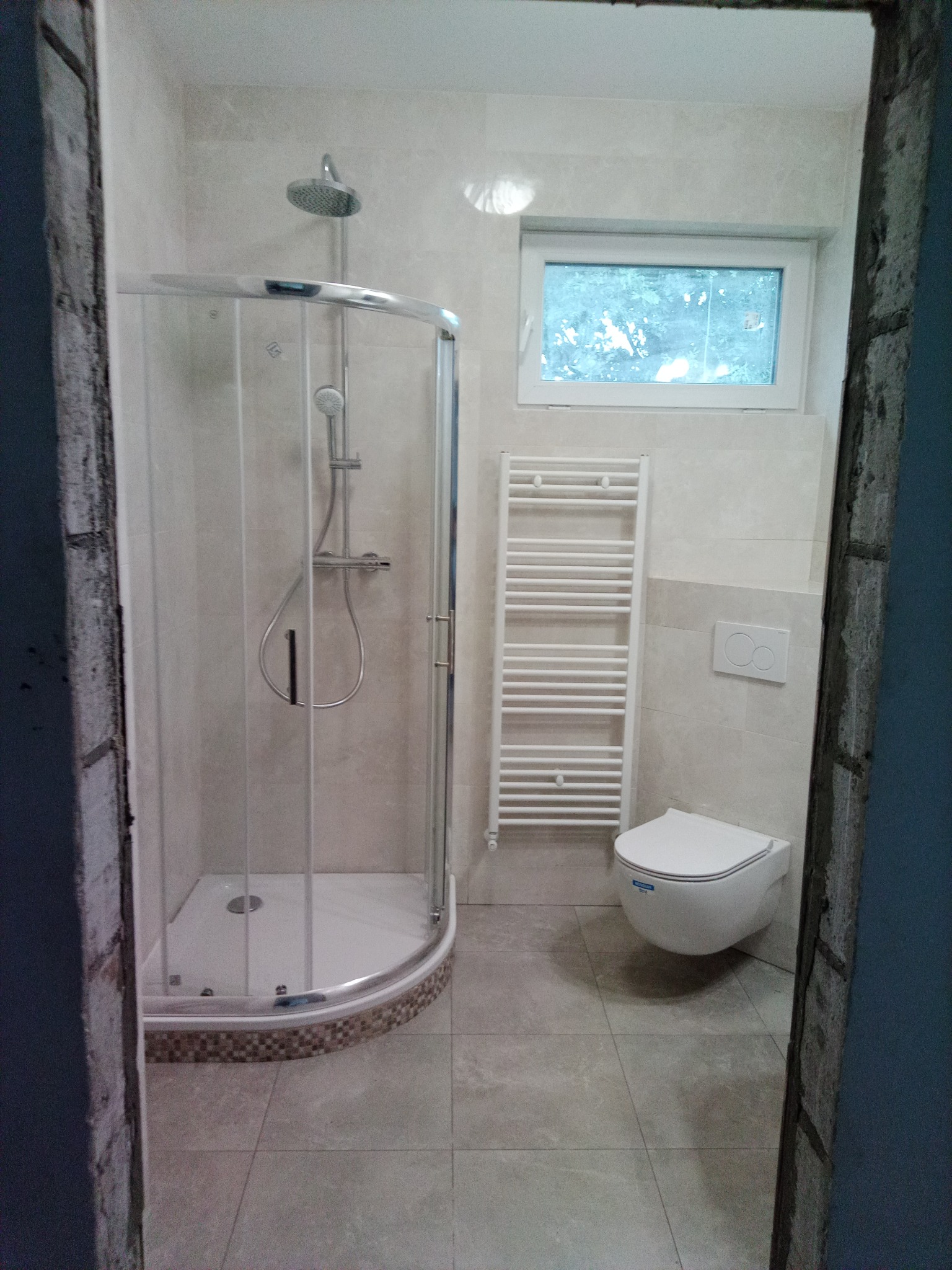 Wykończona łazienka z kabiną prysznicową narożną z mozaiką na brodziku, wiszącą toaletą, grzejnikiem drabinkowym i oknem, widok przez surowy otwór drzwiowy.
