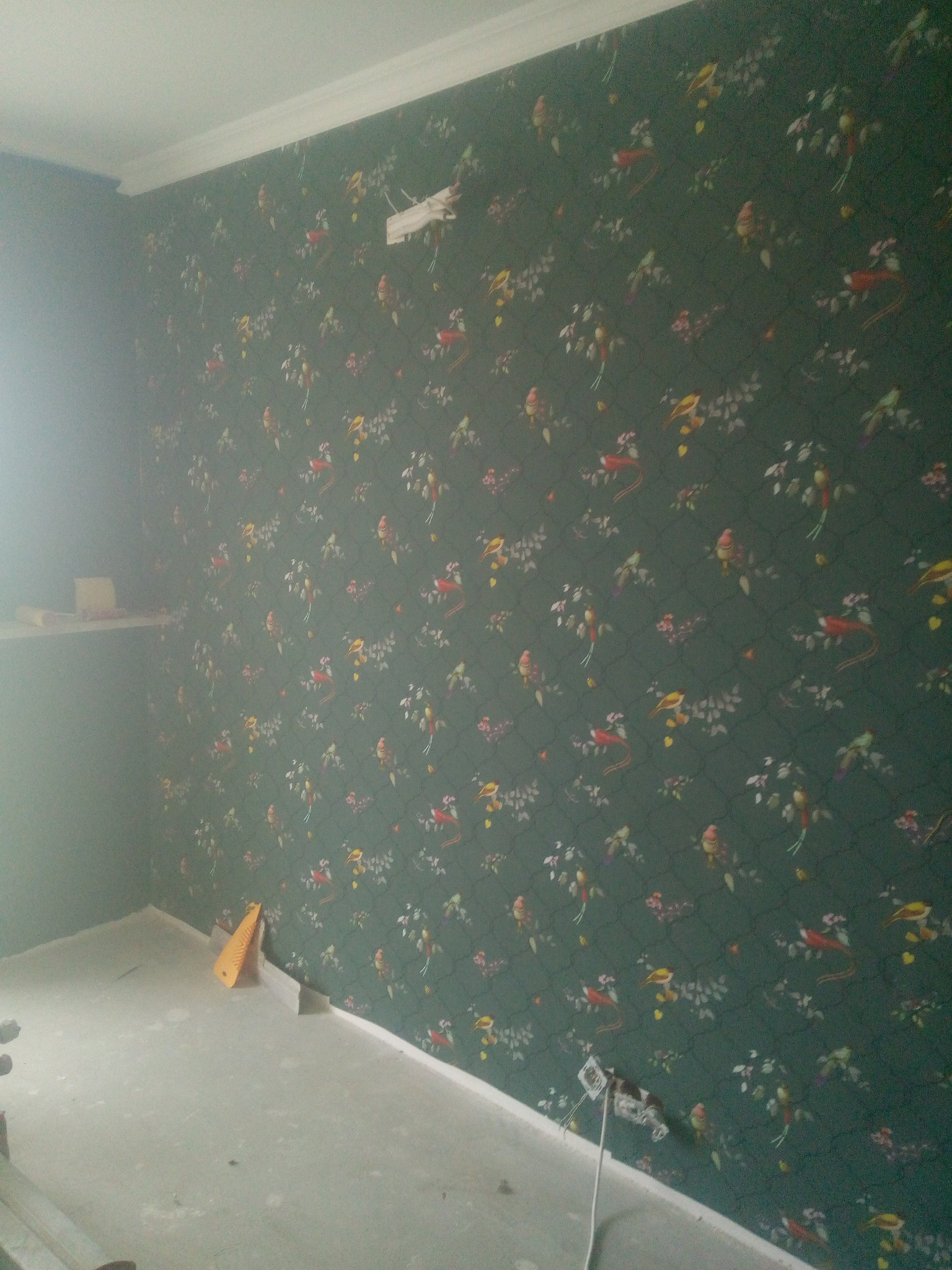 Ściana w pokoju po tapetowaniu, widoczny wzór w ptaki i kwiaty na ciemnozielonym tle, fragment sufitu z listwą dekoracyjną, gniazdko elektryczne z wystającymi kablami, żółta szpachelka oparta...