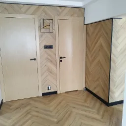 Wykończone wnętrze z dwiema drewnianymi drzwiami i ścianą pokrytą panelami w jodełkę, dopasowanymi do podłogi w tym samym wzorze, z czarnymi akcentami w postaci listew i ramy zegara.