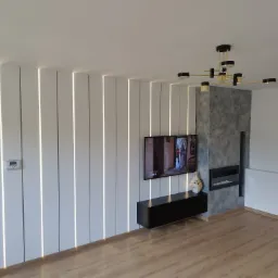 Ściana z płyty karton-gips z pionowymi wnękami oświetlonymi LED, podwieszana szafka RTV i dekoracyjny kominek elektryczny w nowoczesnym salonie.