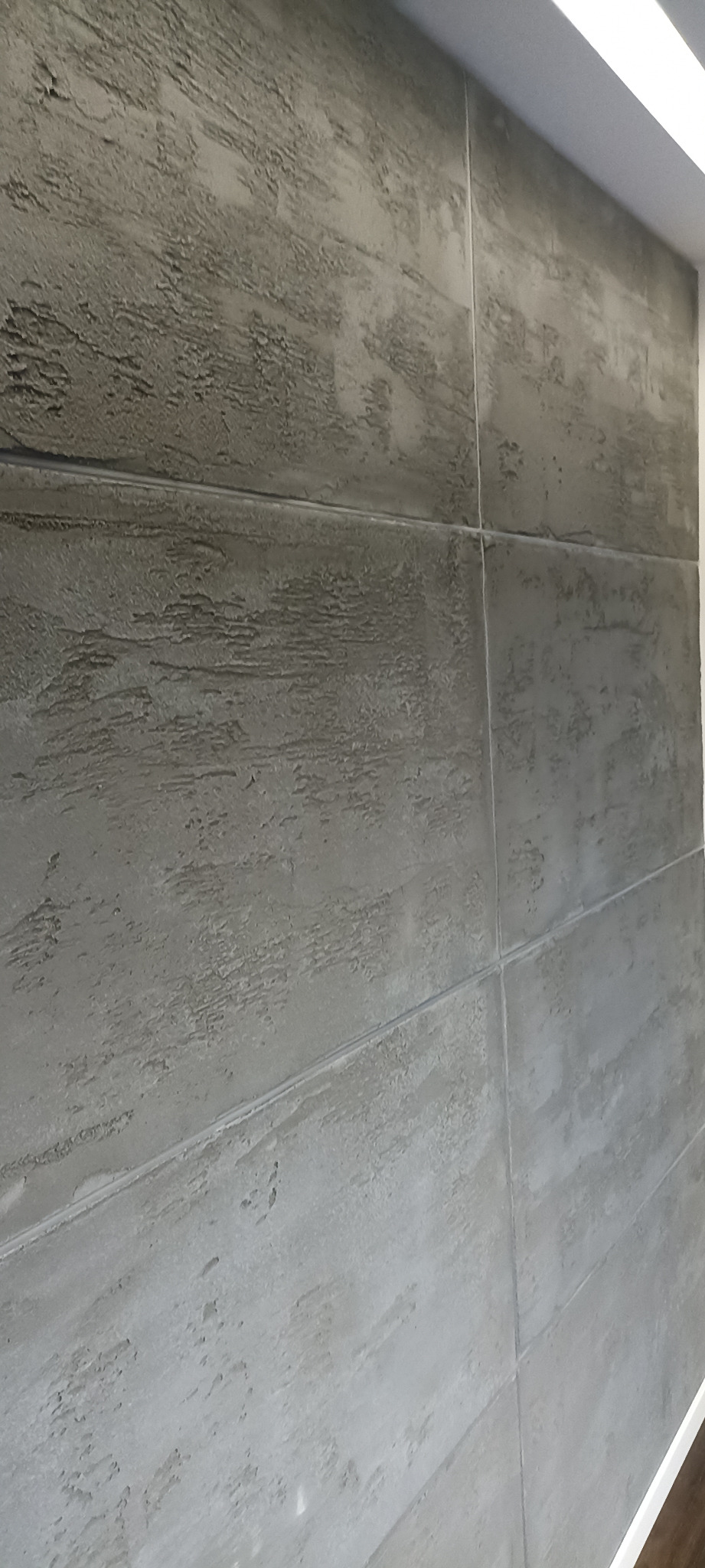 Szara ściana wykończona w panelach imitujących beton z wyraźną, nieregularną fakturą, widoczne łączenia paneli, oświetlenie górne.
