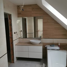 Profesjonalne wykańczanie wnętrz - Łazienka w trakcie remontu na poddaszu: drewnopodobne płytki na ścianach, biała umywalka nablatowa i szafka pod umywalką, widoczne przewody elektryczne wystające z sufitu, fragment wanny...