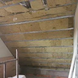 Montaż izolacji z wełny mineralnej w skośnym dachu poddasza, widoczne metalowe profile konstrukcyjne i fragment rusztowania z boku.