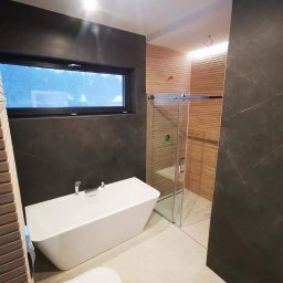 Profesjonalne wykańczanie wnętrz - Nowoczesna łazienka z wolnostojącą białą wanną, kabiną prysznicową ze szklanymi drzwiami, toaletą i płytkami imitującymi drewno na ścianie oraz ciemnymi płytkami na pozostałych ścianach.