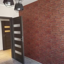 Profesjonalne wykańczanie wnętrz - Ściana z tapetą imitującą czerwoną cegłę w nowoczesnym wnętrzu, widoczne otwarte drzwi wewnętrzne z przeszkleniami, szare panele podłogowe i czarne, geometryczne lampy sufitowe.