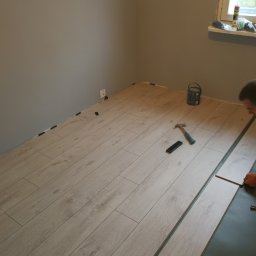Profesjonalne wykańczanie wnętrz - Fragment pokoju w trakcie montażu paneli podłogowych w odcieniach jasnego drewna; widoczne narzędzia: młotek, listwa dystansowa, oraz puszka farby przy oknie.
