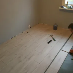 Fragment pokoju w trakcie montażu paneli podłogowych w odcieniach jasnego drewna; widoczne narzędzia: młotek, listwa dystansowa, oraz puszka farby przy oknie.