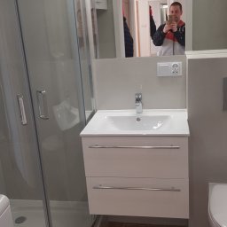 Profesjonalne wykańczanie wnętrz - Nowoczesna łazienka po remoncie: kabina prysznicowa z zaokrąglonym szkłem, biała szafka z umywalką i chromowaną armaturą, drewnopodobne płytki podłogowe, widoczne odbicie fotografa w lustrze.