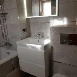 Wykończona łazienka z szarymi płytkami imitującymi fale, białą umywalką z szafką, podświetlanym lustrem i chromowaną armaturą natryskową.