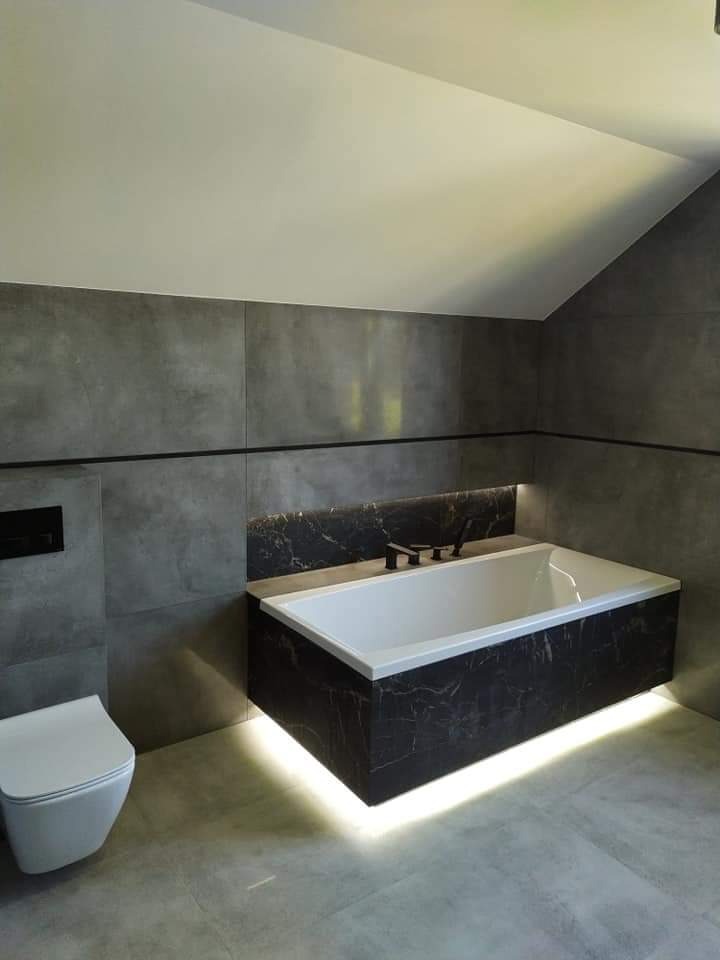 Nowoczesna łazienka w szarościach z podświetlaną wanną obłożoną czarnym marmurem i wiszącą toaletą.