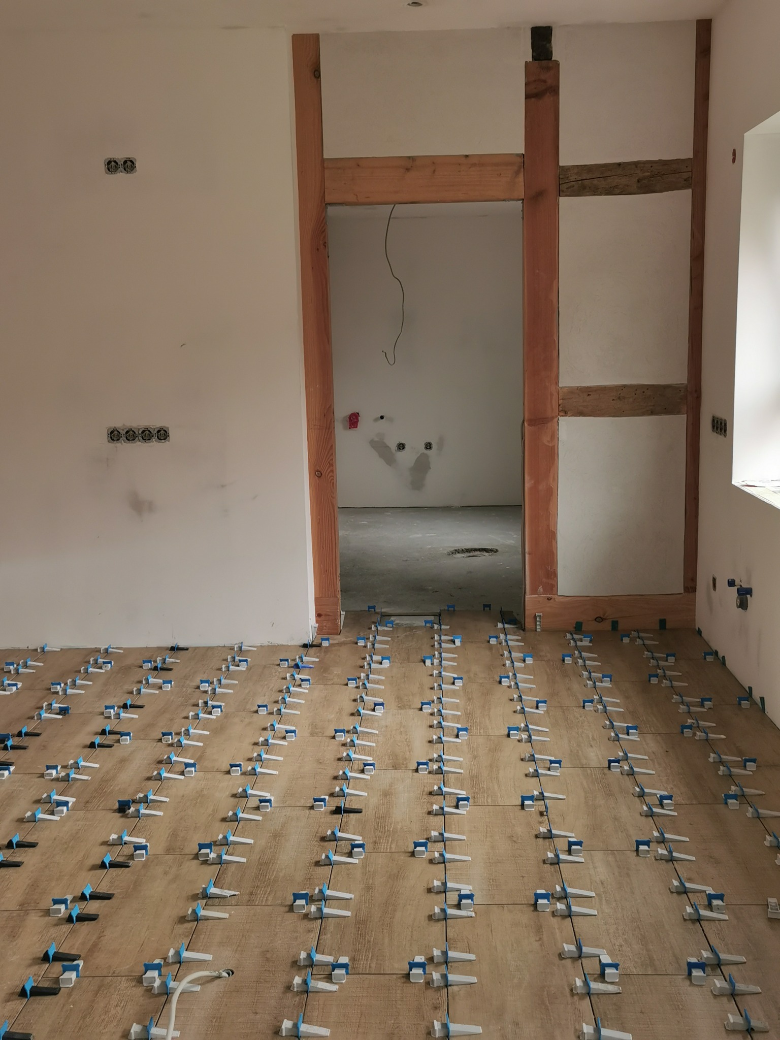 Ułożone płytki podłogowe z systemem poziomowania, widoczne drewniane elementy konstrukcyjne w tle, ściany przygotowane do dalszych prac wykończeniowych, gniazdka elektryczne na ścianie.