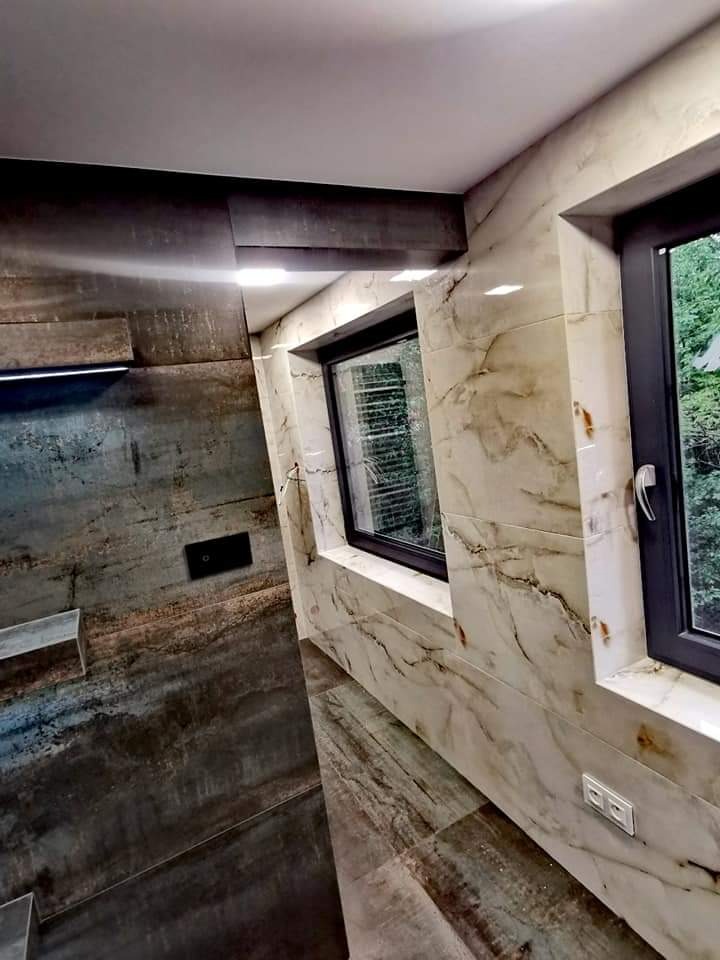 Wykończona łazienka z marmurowymi płytkami na ścianach i podłodze, dwa okna z czarnymi ramami, oświetlenie punktowe i ukryta spłuczka w szarej ścianie.