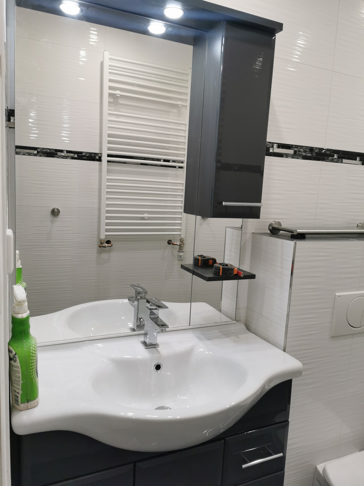 Nowoczesna łazienka po remoncie: białe płytki z dekoracyjnym pasem, umywalka w szafce z szufladami w kolorze grafitowym, grzejnik drabinkowy i wisząca szafka z półką, na której leżą miarki.