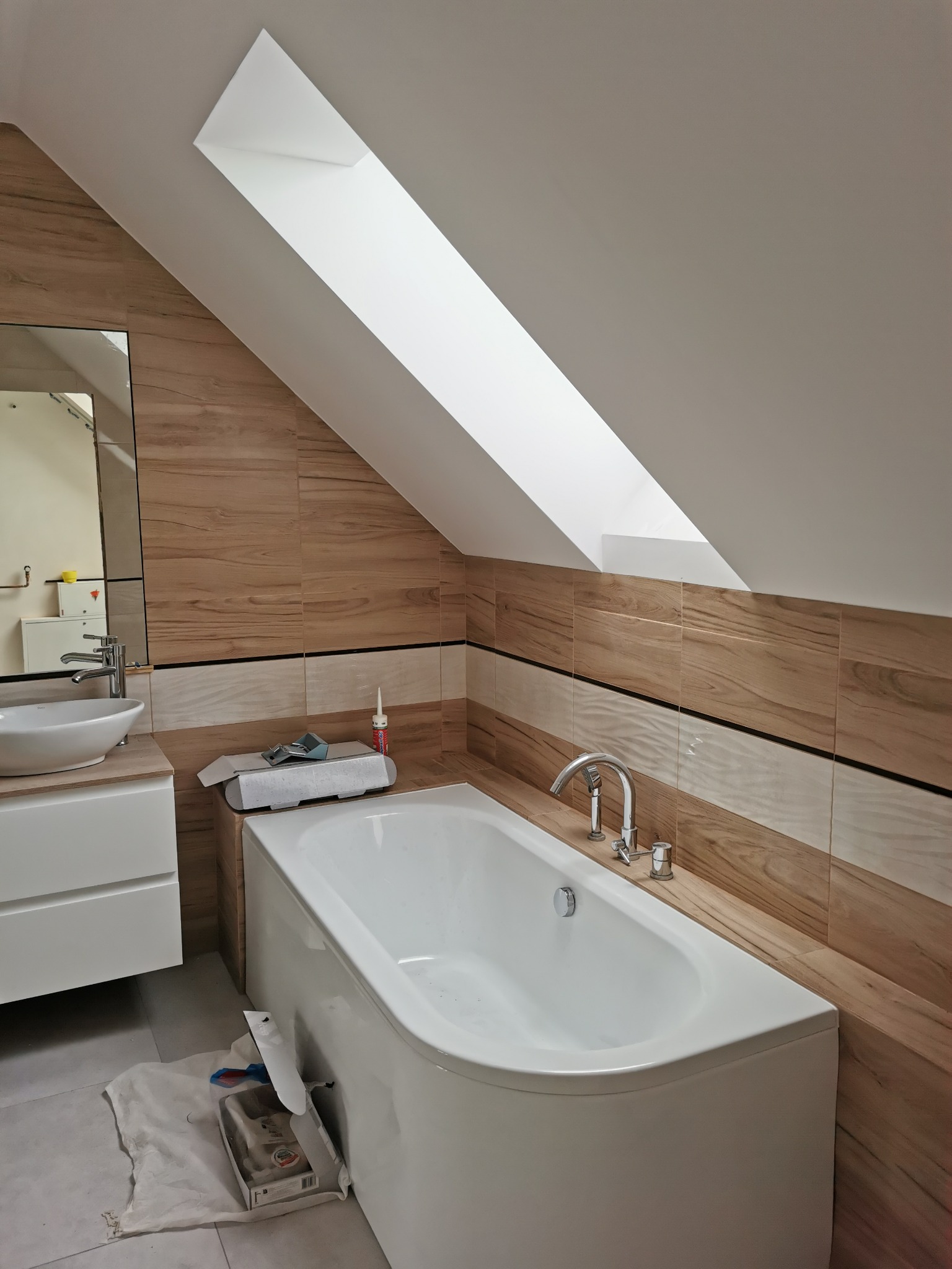 Biała wanna narożna zamontowana pod skosem dachu z oknem, obłożona płytkami imitującymi drewno, widoczna umywalka nablatowa i akcesoria glazurnicze.