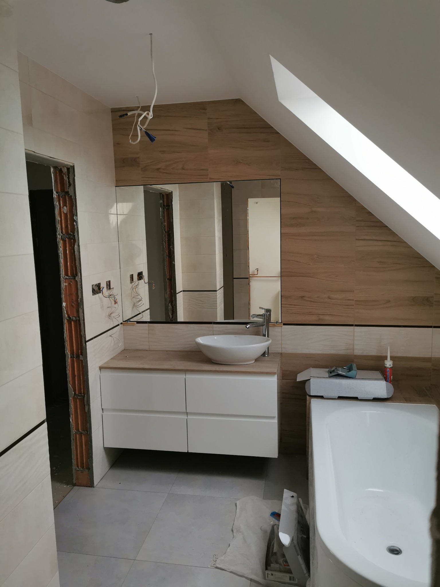 Łazienka w trakcie remontu na poddaszu: drewnopodobne płytki na ścianach, biała umywalka nablatowa i szafka pod umywalką, widoczne przewody elektryczne wystające z sufitu, fragment wanny...