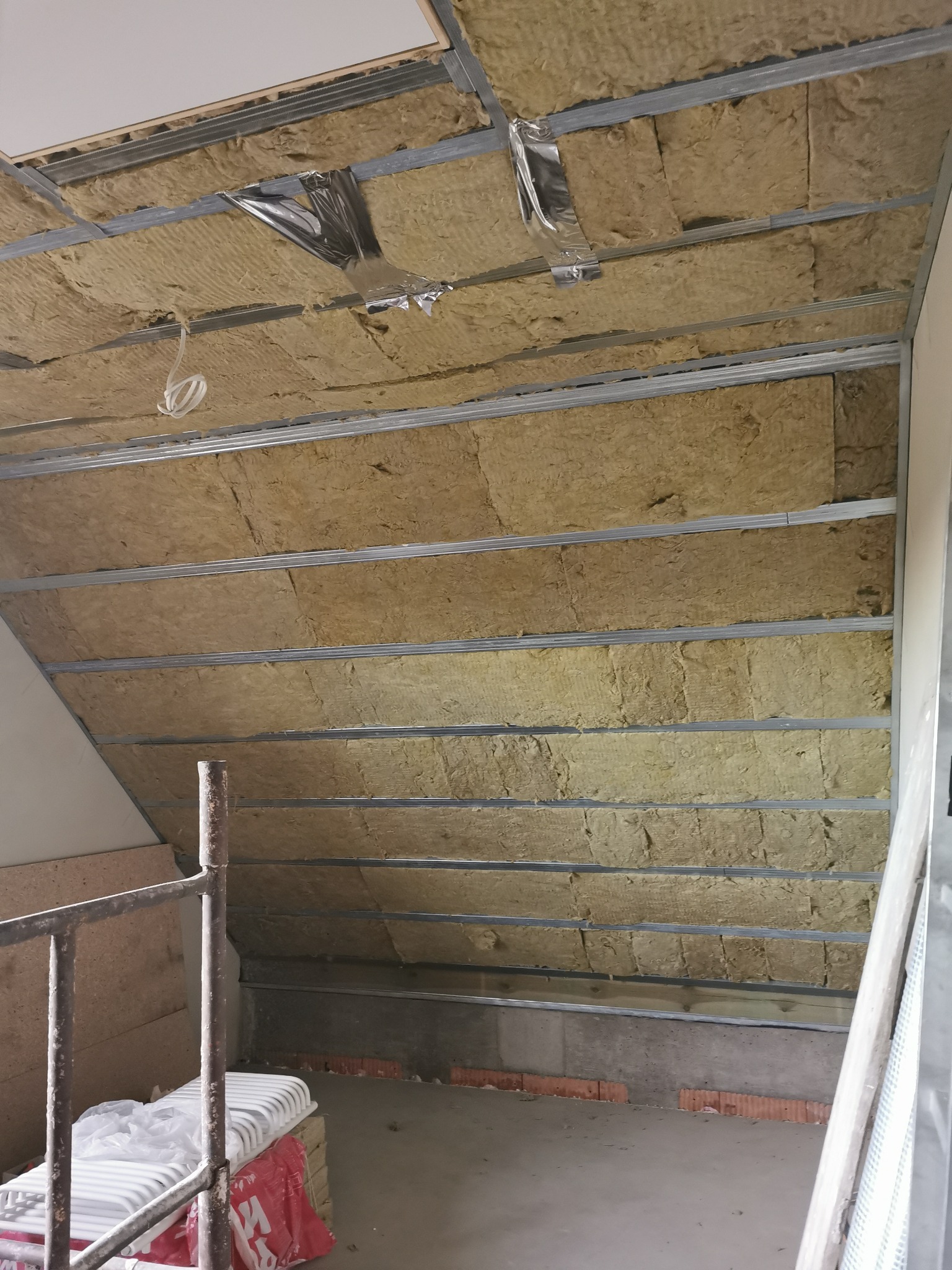 Montaż izolacji z wełny mineralnej w skośnym dachu poddasza, widoczne metalowe profile konstrukcyjne i fragment rusztowania z boku.