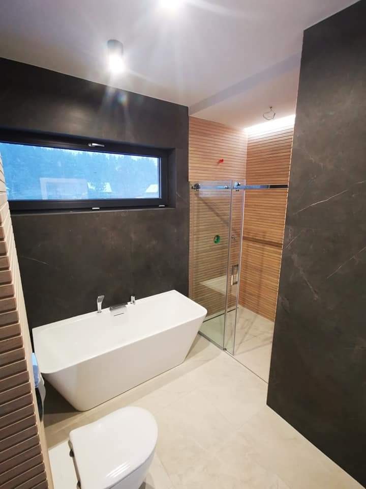 Nowoczesna łazienka z wolnostojącą białą wanną, kabiną prysznicową ze szklanymi drzwiami, toaletą i płytkami imitującymi drewno na ścianie oraz ciemnymi płytkami na pozostałych ścianach.