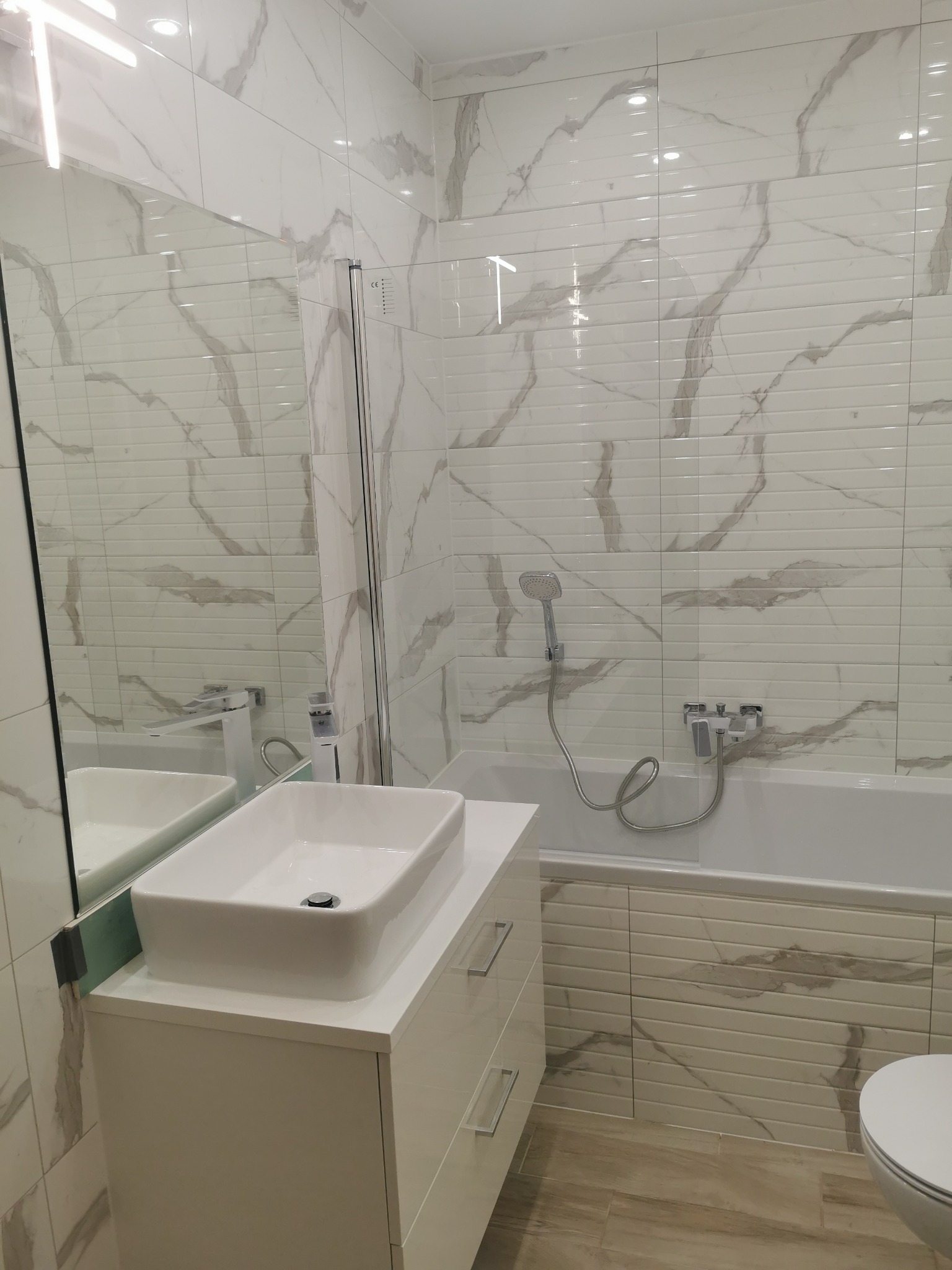 Nowoczesna łazienka z białymi płytkami imitującymi marmur, wisząca umywalka na szafce z szufladami oraz widoczna wanna z baterią i prysznicem.