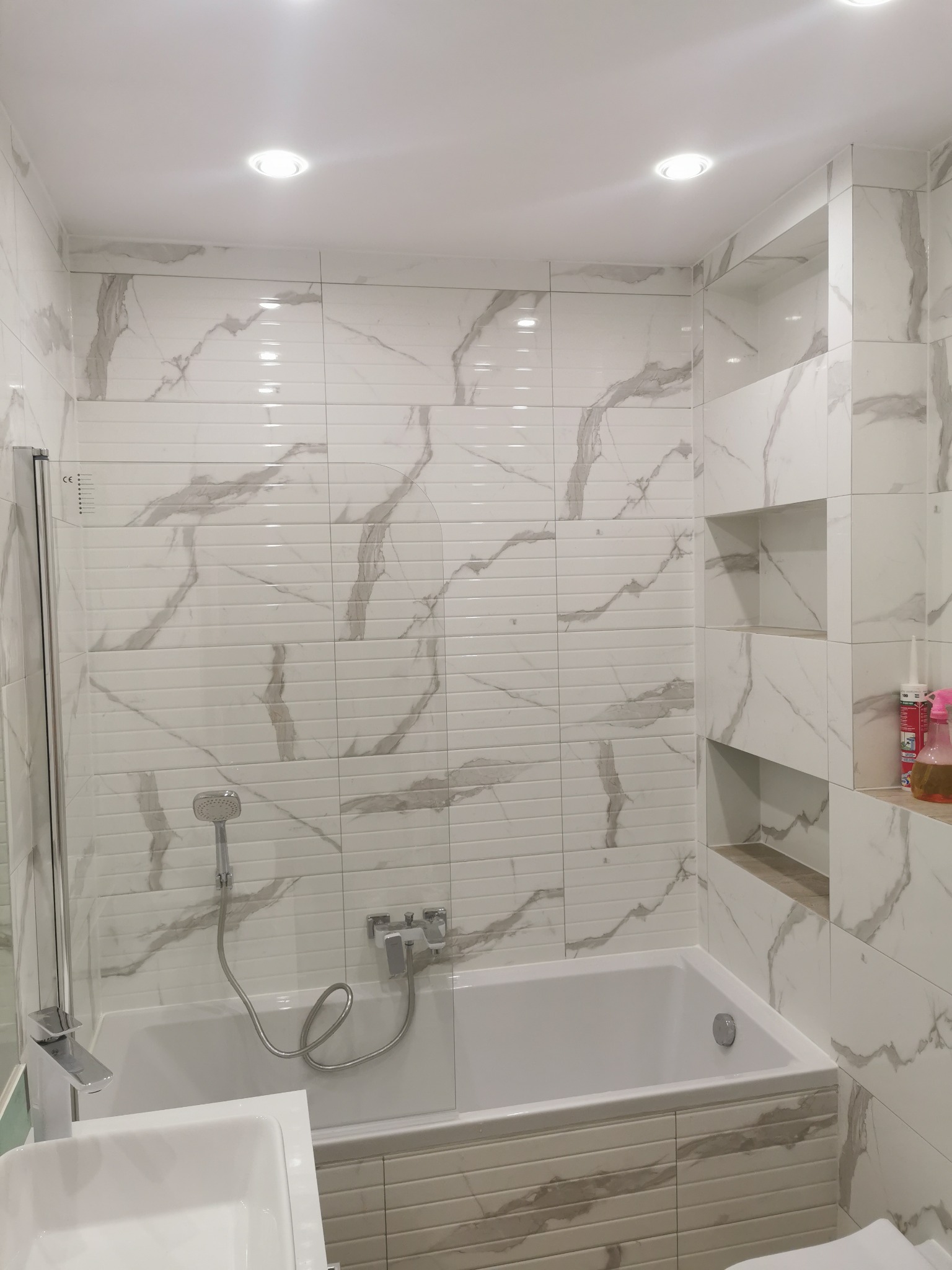 Wykończona łazienka z wanną, umywalką i kabiną prysznicową, wyłożona białymi płytkami imitującymi marmur z szarymi żyłami, wnęki w ścianie wykończone płytkami w odcieniach szarości.
