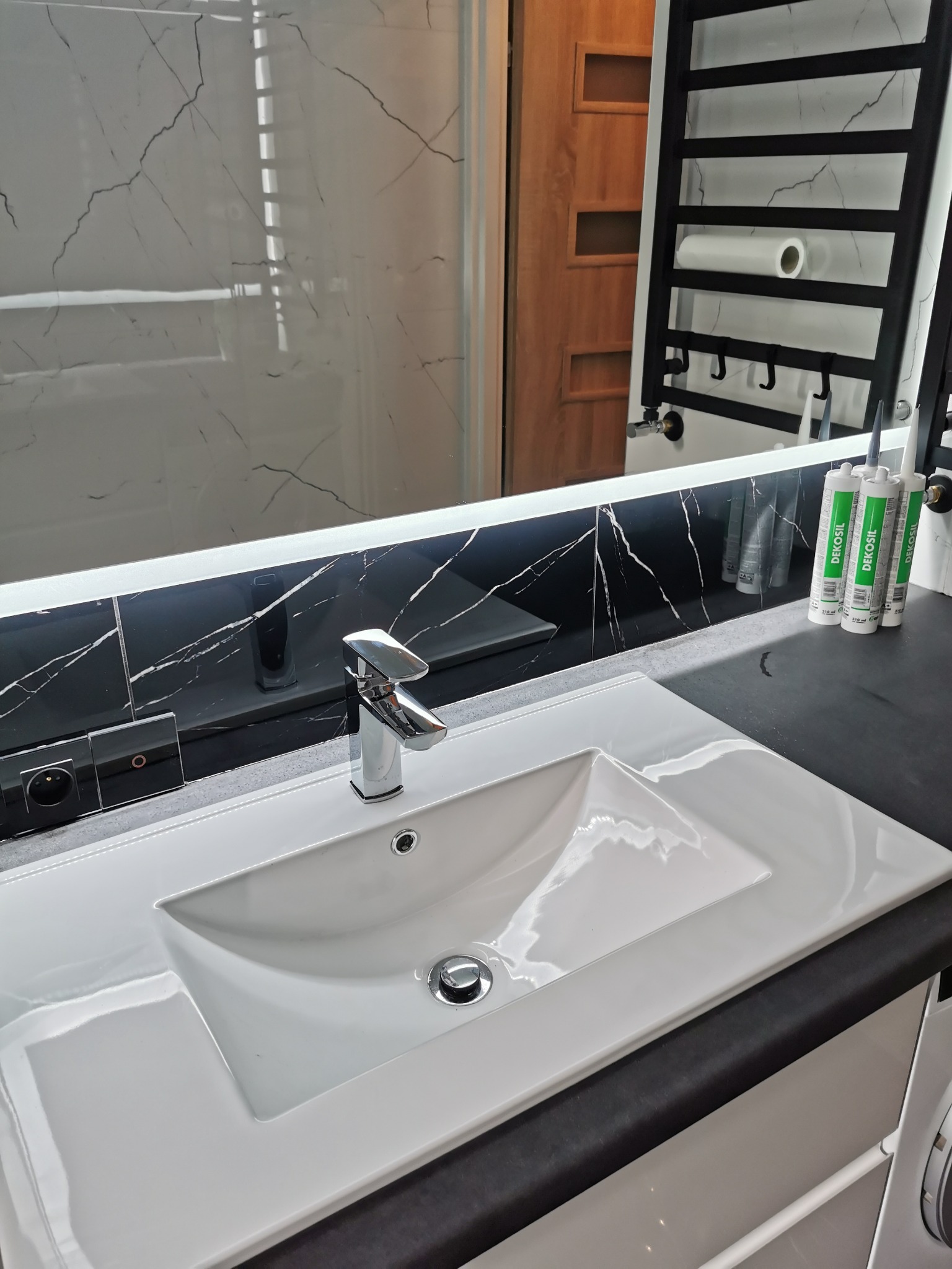 Nowoczesna łazienka z białą umywalką wpuszczaną w czarny blat, chromowaną baterią, podświetlanym lustrem z czarnym marmurowym wzorem, grzejnikiem drabinkowym i tubami silikonu DEKOSIL.