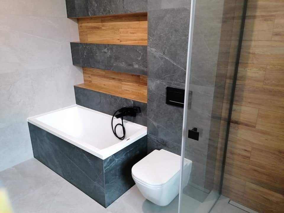Nowoczesna łazienka z wanną obudowaną płytkami imitującymi kamień, toaletą wiszącą, kabiną prysznicową z drewnopodobną ścianą i dekoracyjnymi półkami z szarego kamienia i drewna.