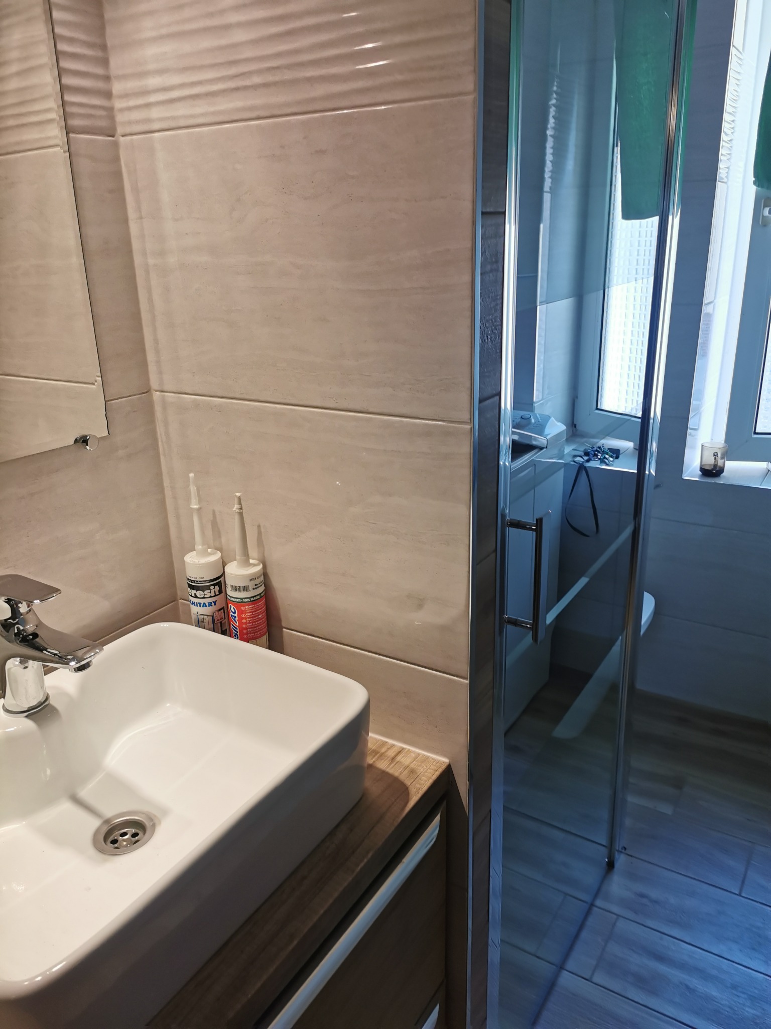 Wykończona łazienka z umywalką nablatową, kabiną prysznicową z przezroczystego szkła oraz kremowymi płytkami na ścianach. Widoczne tuby z silikonem sanitarnym marki Ceresit.