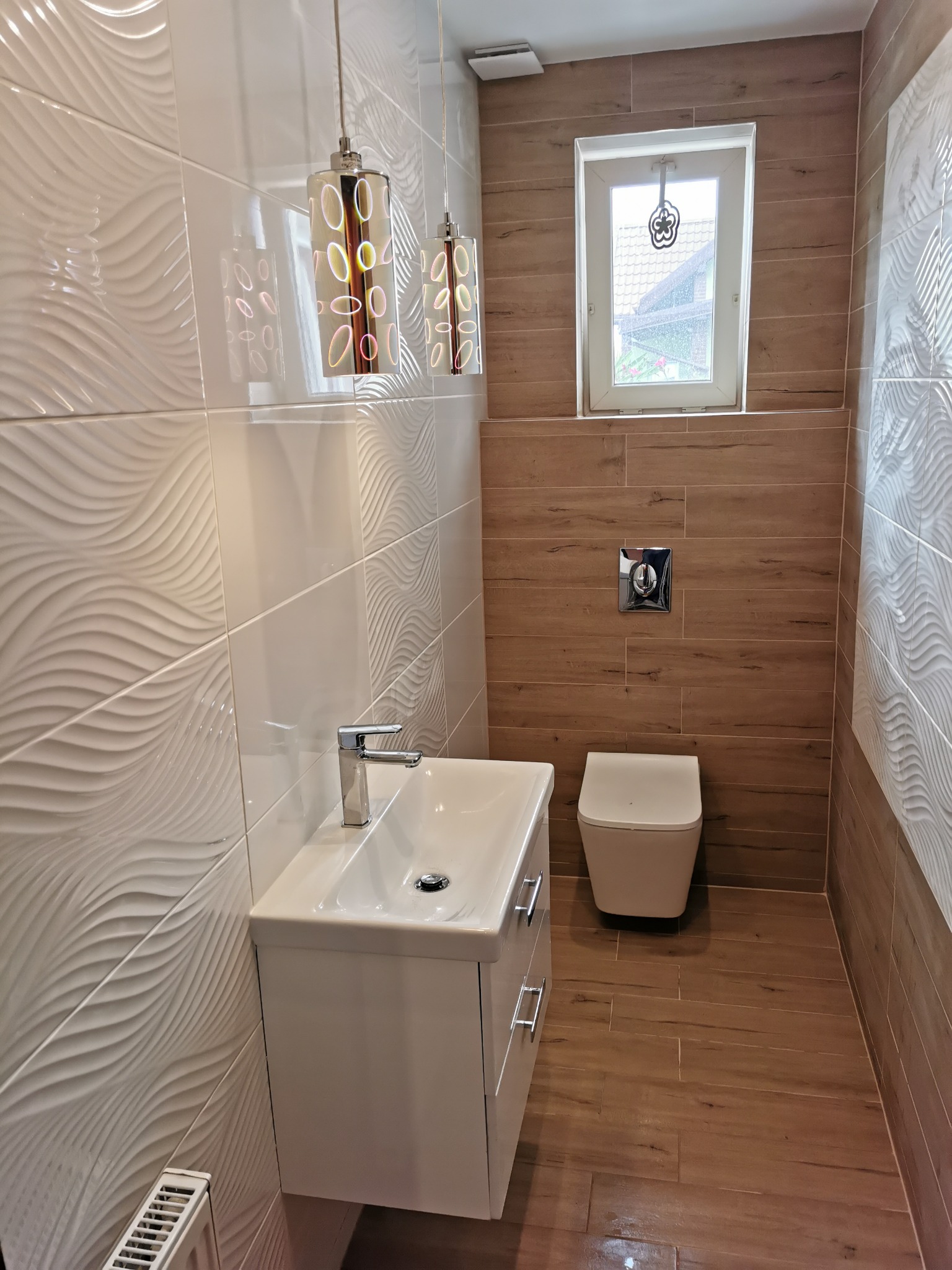 Wykończona łazienka z białymi, trójwymiarowymi płytkami ściennymi, brązowymi płytkami imitującymi drewno, wiszącą szafką z umywalką, wiszącą toaletą i dekoracyjnymi lampami wiszącymi.