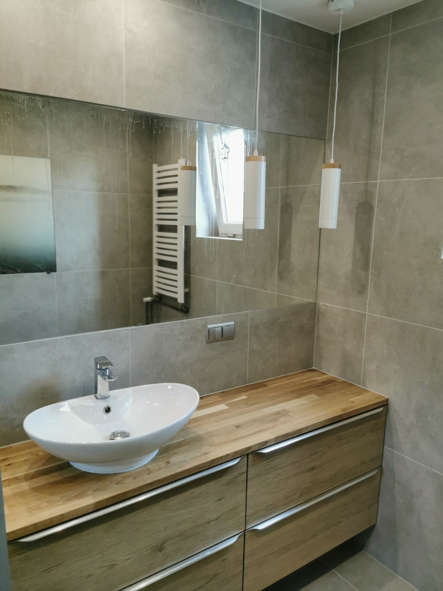 Nowoczesna łazienka z szarą płytką, drewnianym blatem pod umywalką i wiszącymi lampami z drewnianymi akcentami, widoczne krople wody na lustrze.