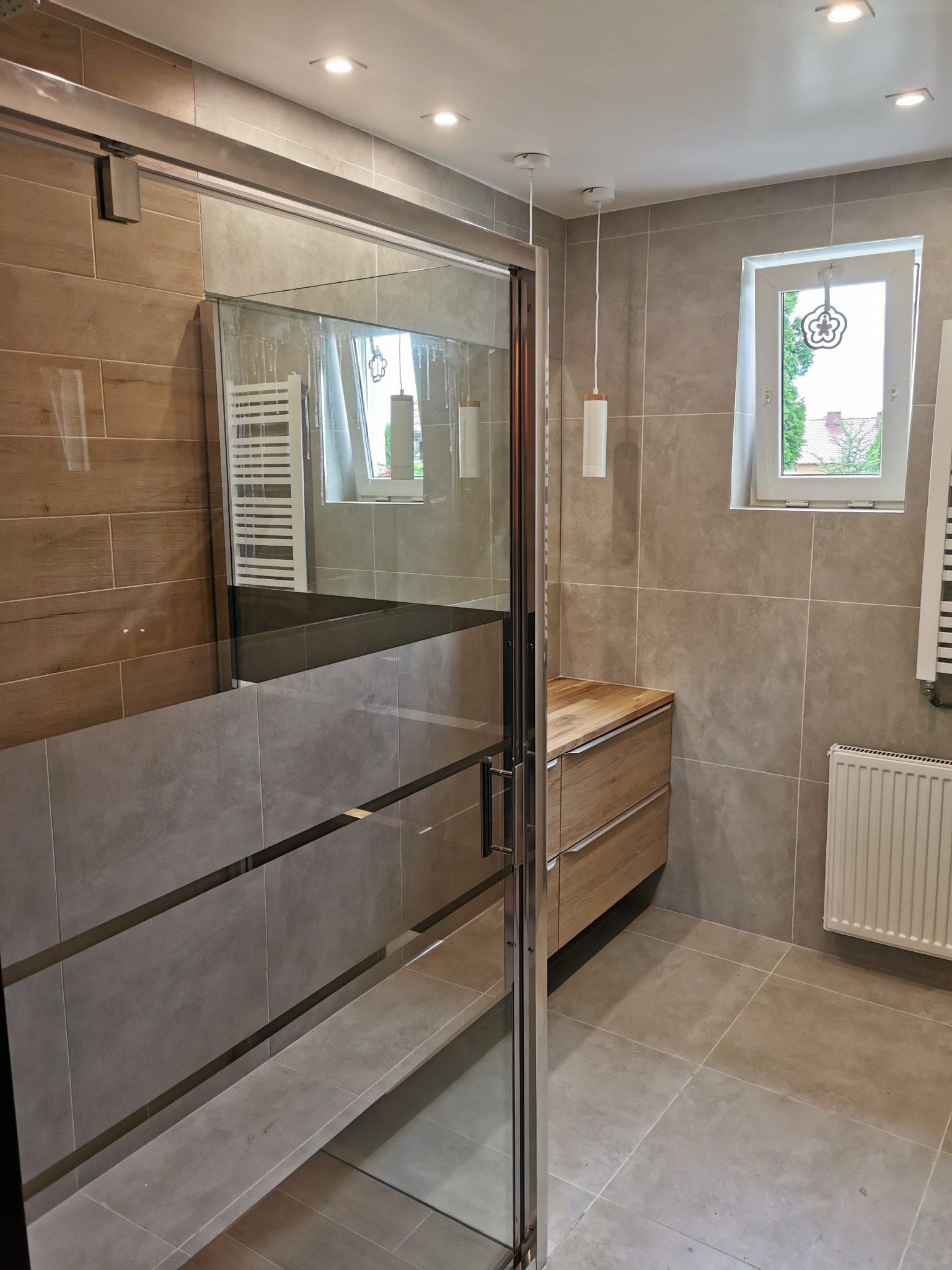 Nowoczesna łazienka z przesuwną szklaną kabiną prysznicową, szare płytki imitujące kamień na ścianach i podłodze, drewniana szafka pod umywalką z dwoma szufladami, grzejnik drabinkowy i okno...