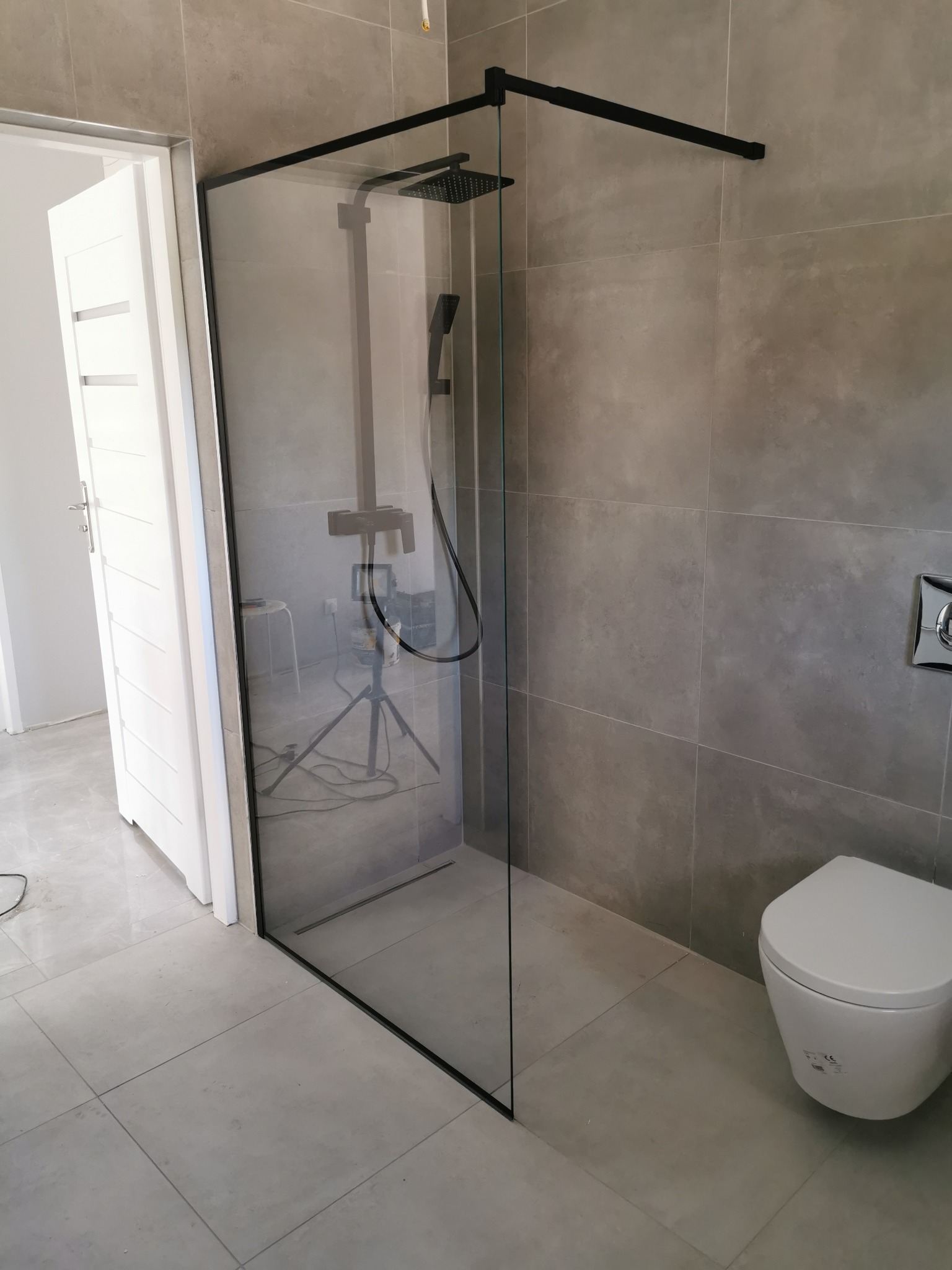 Nowoczesna kabina prysznicowa z czarnymi ramami i deszczownicą, szare płytki na ścianach, widoczna toaleta podwieszana i białe drzwi w tle, odbicie aparatu na statywie w szkle.