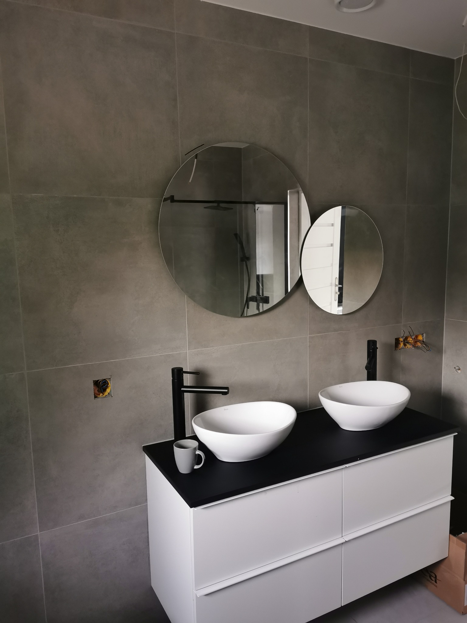 Łazienka z szarymi płytkami, dwiema okrągłymi umywalkami na białej szafce z czarnym blatem, dwoma okrągłymi lustrami i widocznymi puszkami elektrycznymi w ścianie.
