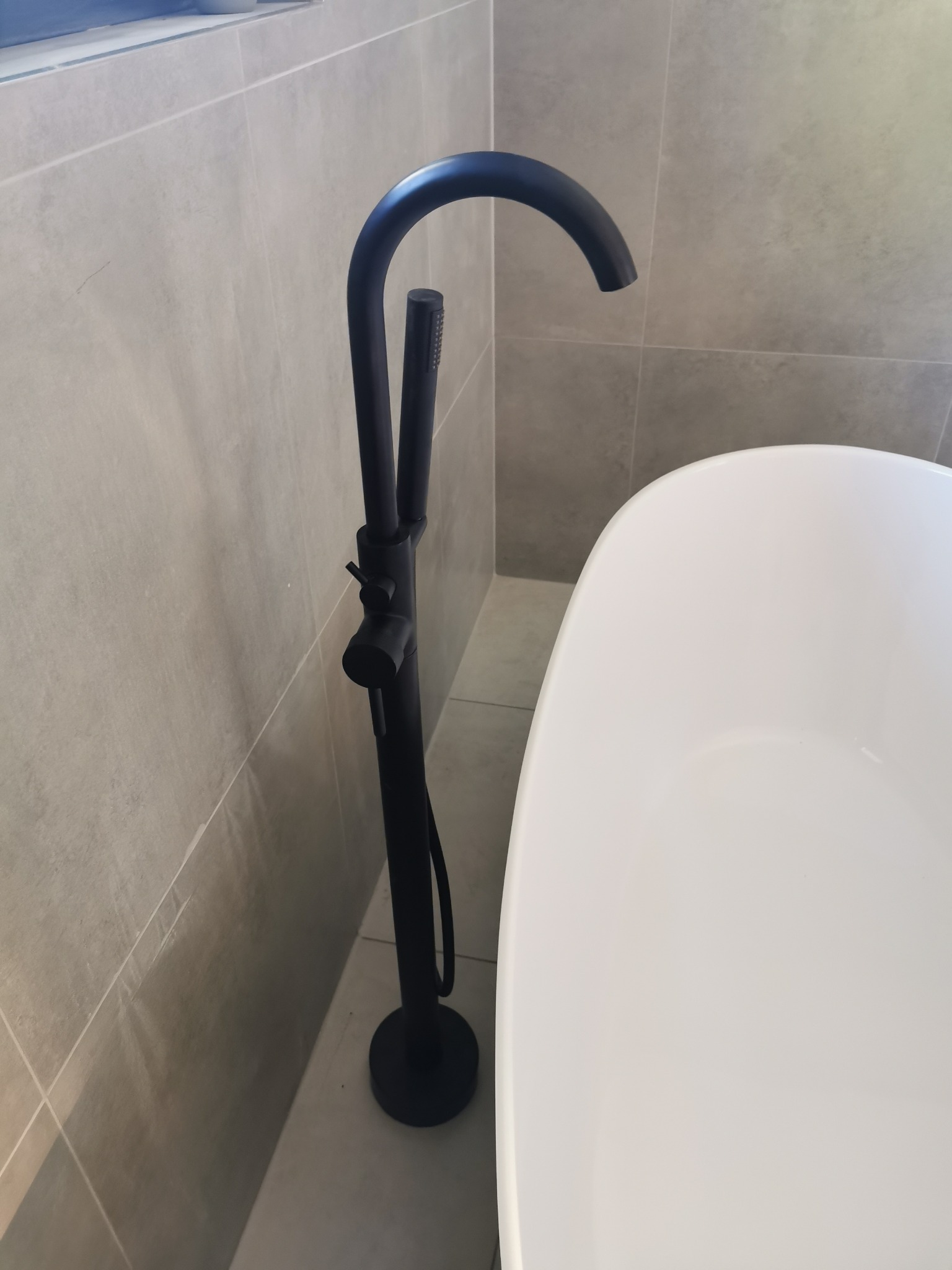 Wolnostojąca, czarna bateria wannowa z słuchawką prysznicową, zamontowana obok białej wanny na tle szarych płytek.