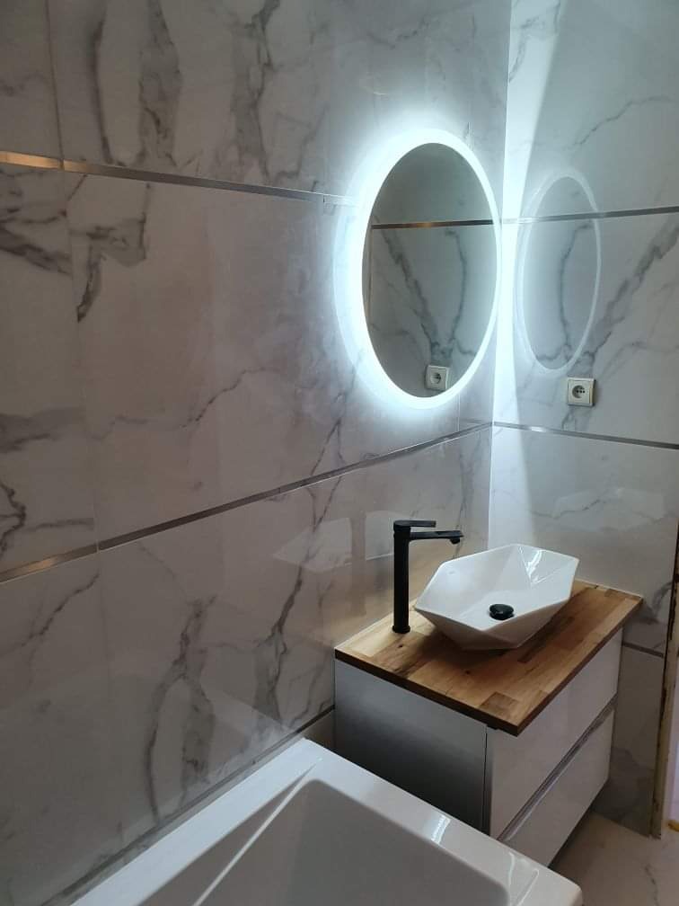 Wykończona łazienka z marmurowymi płytkami, okrągłym lustrem z podświetleniem LED, białą umywalką o geometrycznym kształcie i czarną armaturą na drewnianym blacie.
