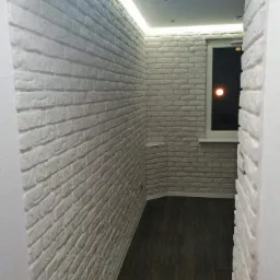 Wąskie pomieszczenie wykończone dekoracyjną białą cegłą, oświetlone listwą LED pod sufitem i z ciemną podłogą z płytek, widok na nocne niebo przez okno.