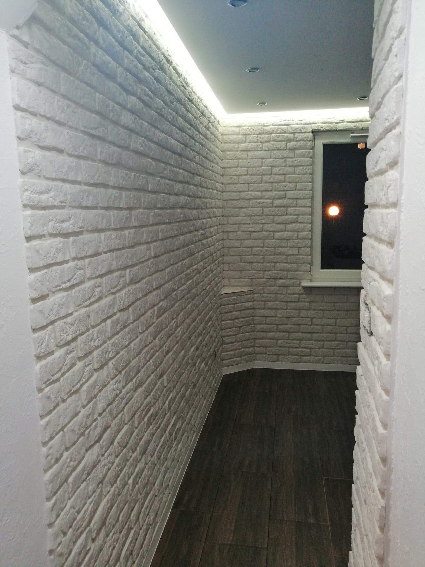 Wąskie pomieszczenie wykończone dekoracyjną białą cegłą, oświetlone listwą LED pod sufitem i z ciemną podłogą z płytek, widok na nocne niebo przez okno.