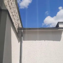 Widok z okna na fragment elewacji budynku z kamienną fakturą, rynnę i błękitne niebo z chmurami, widoczne smugi na szybie.