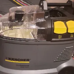 Profesjonalny odkurzacz piorący Kärcher Puzzi 8/1 C z przezroczystym zbiornikiem na wodę i żółtymi przyciskami, leżący na łóżku w kolorowe paski, z widoczną końcówką do prania.