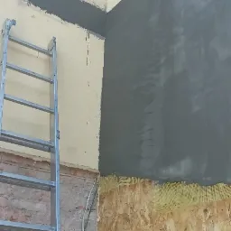 Fragment elewacji budynku w trakcie prac: ocieplenie płyty OSB z siatką i częściowo nałożona warstwa szarego tynku, widoczna drabina aluminiowa i fragment okna na górze.