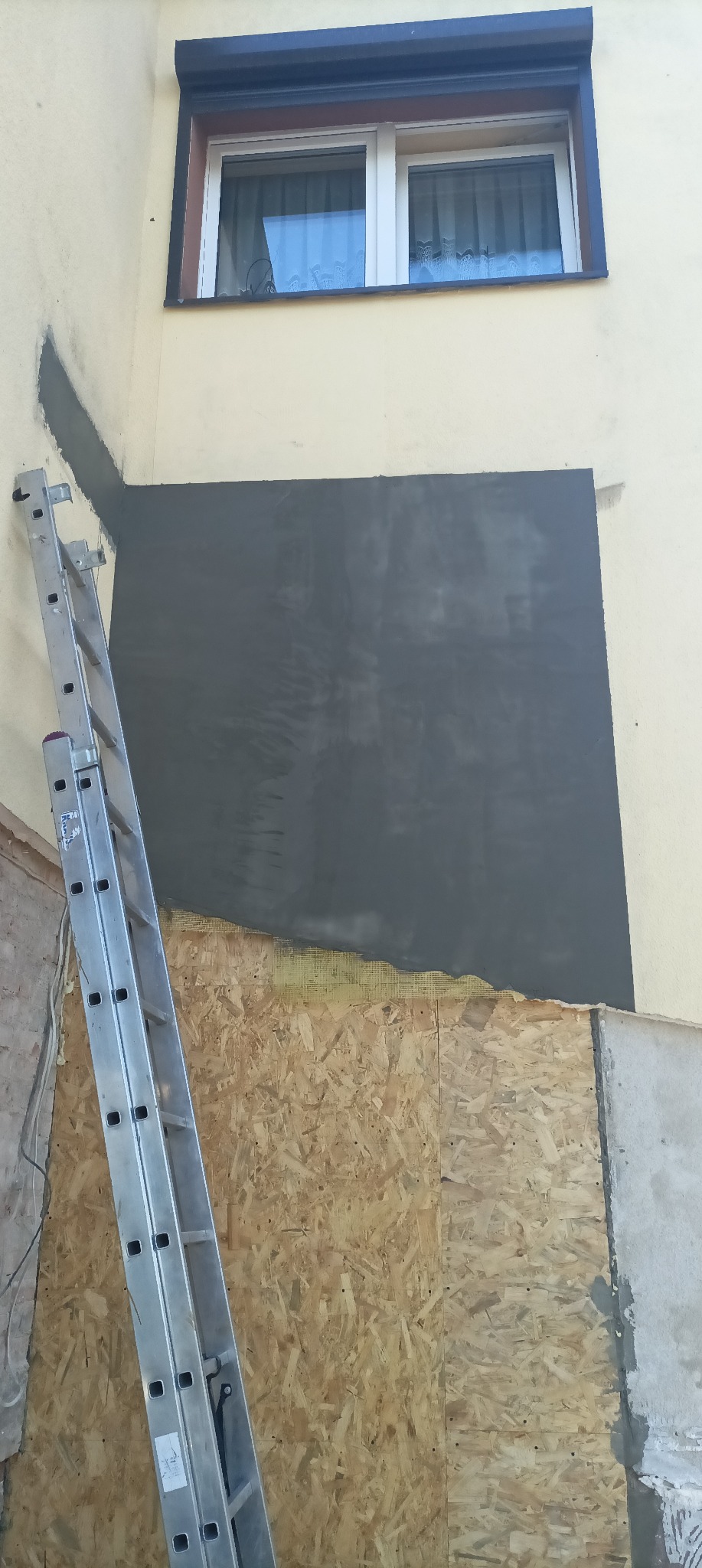 Zamurowane okno z widoczną płytą OSB, siatką i warstwą szarego tynku, z drabiny opartą o ścianę.