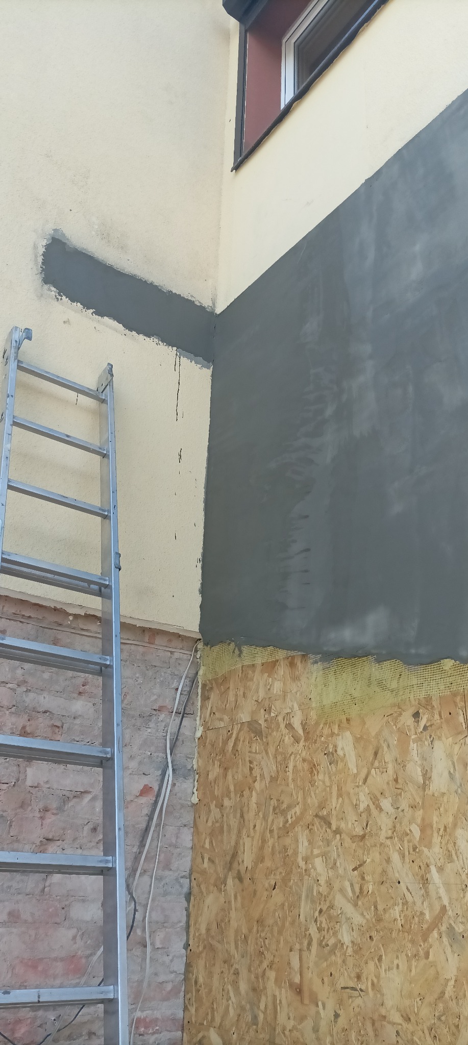 Fragment elewacji budynku w trakcie prac: ocieplenie płyty OSB z siatką i częściowo nałożona warstwa szarego tynku, widoczna drabina aluminiowa i fragment okna na górze.