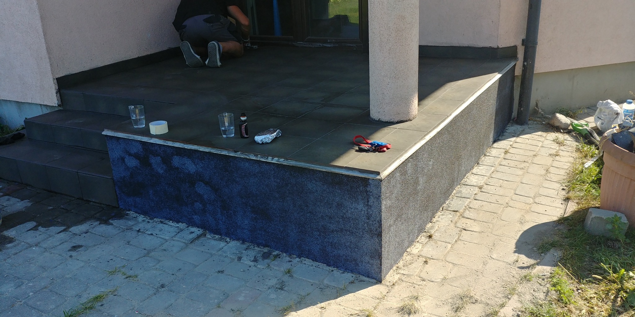 Prace wykończeniowe na tarasie: płytki podłogowe, obłożona ściana, pracownik na kolanach, szklanki z wodą, folia aluminiowa i narzędzia.