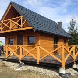 Żółty dom z bali z ciemnoszarym dachem, drewnianą balustradą wokół tarasu i osobą w zielonej kurtce pracującą na tarasie.