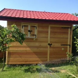 Drewniana konstrukcja z czerwonym dachem, stylizowana na mały domek, z dekoracyjnymi okuciami przy oknie i drzwiach, otoczona zielenią ogrodu i siatką ogrodzeniową.