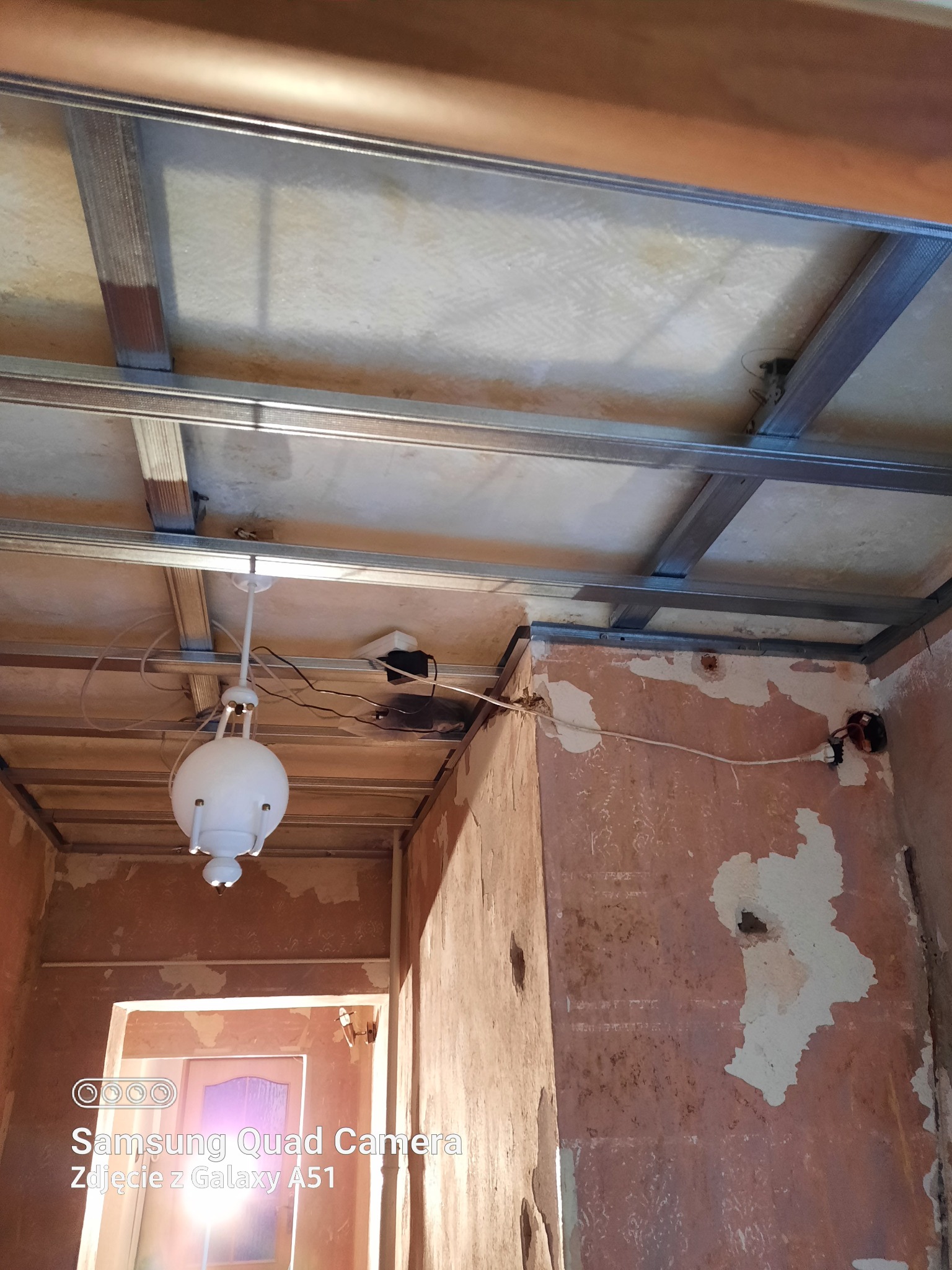 Wnętrze w trakcie remontu: widoczne metalowe profile sufitowe, stara instalacja elektryczna z wiszącą lampą kulą i fragmenty odrapanych ścian.