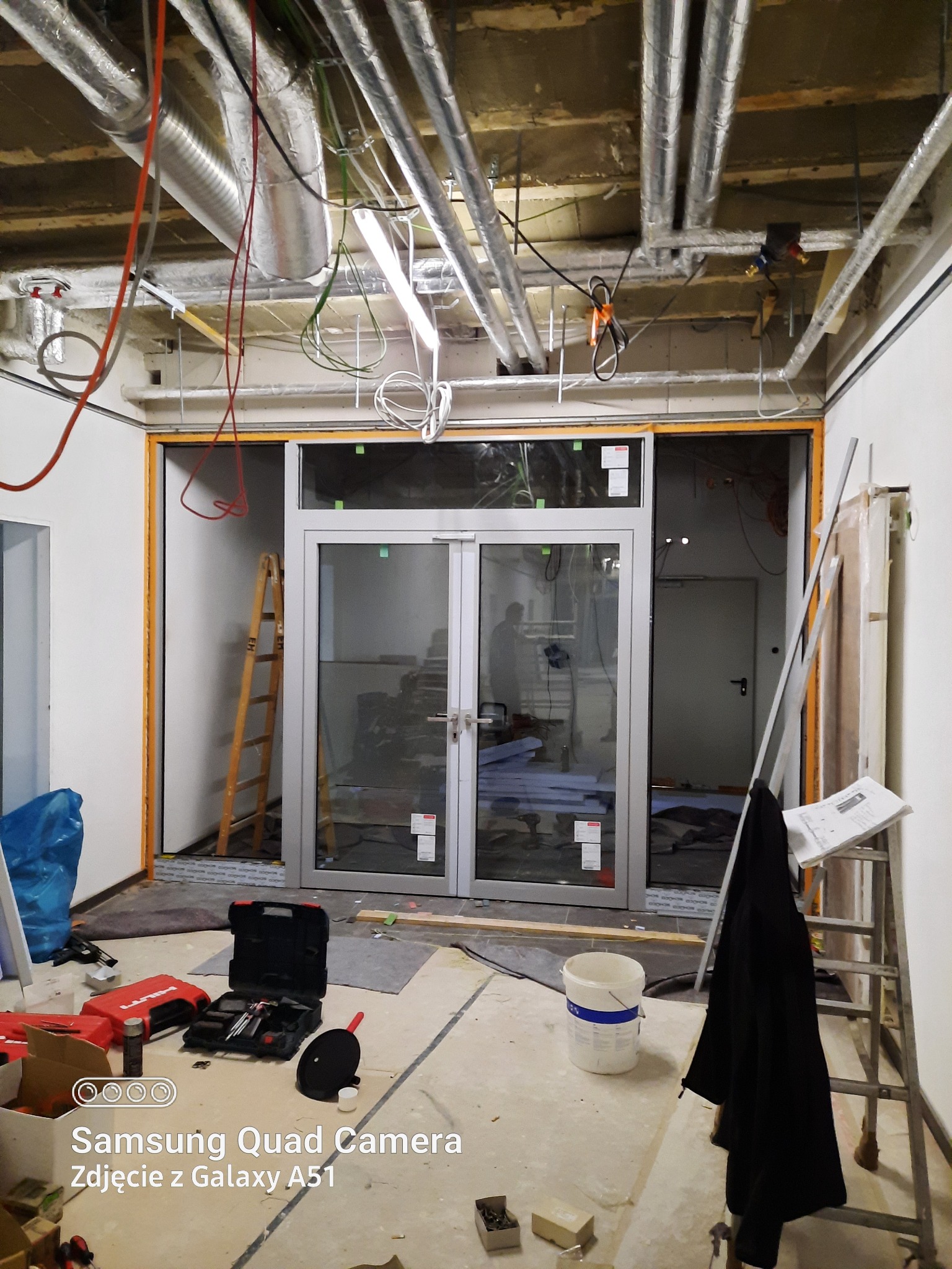 Montaż aluminiowych drzwi dwuskrzydłowych w nowo wykończonym pomieszczeniu, widoczne narzędzia, drabiny, rury wentylacyjne pod sufitem i surowy stan wykończenia.