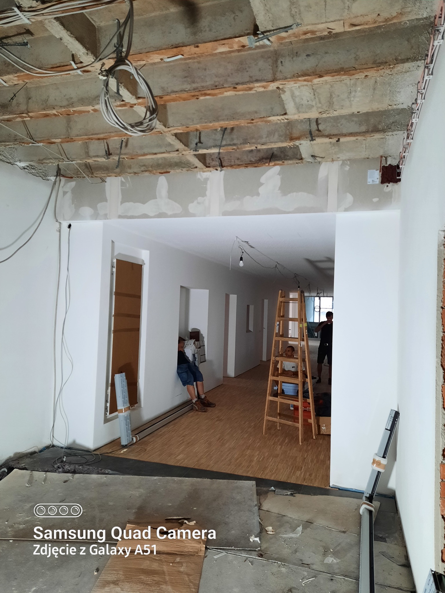 Wnętrze w trakcie remontu w Lublińcu: widoczne przewody elektryczne pod sufitem, pracownik siedzący na podłodze, drabina i przygotowane materiały budowlane.