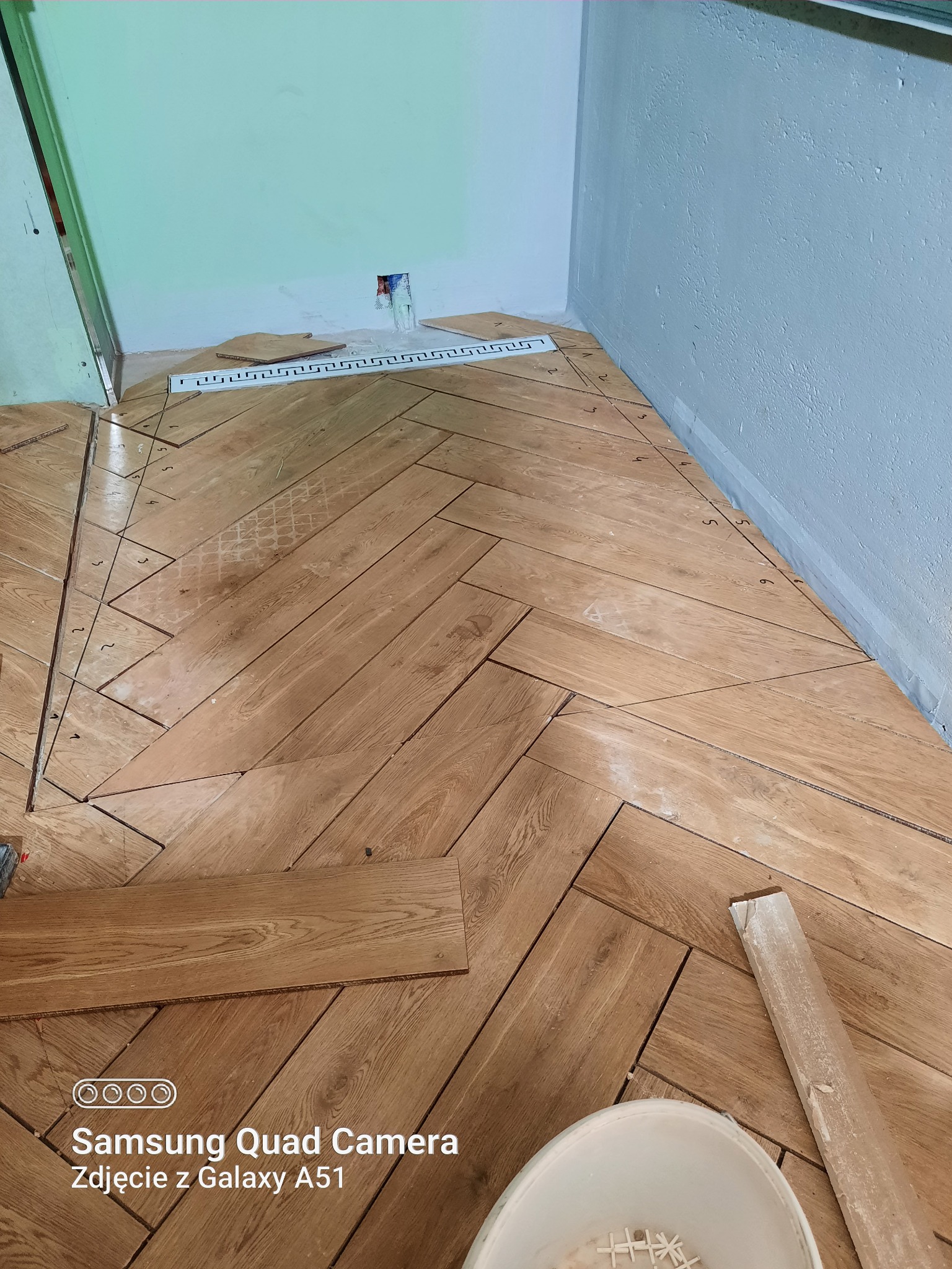 Układanie parkietu w jodełkę z jasnego drewna, widoczne oznaczenia na podłodze, biała listwa przypodłogowa i wiadro z krzyżykami dystansowymi w narożniku pomieszczenia.