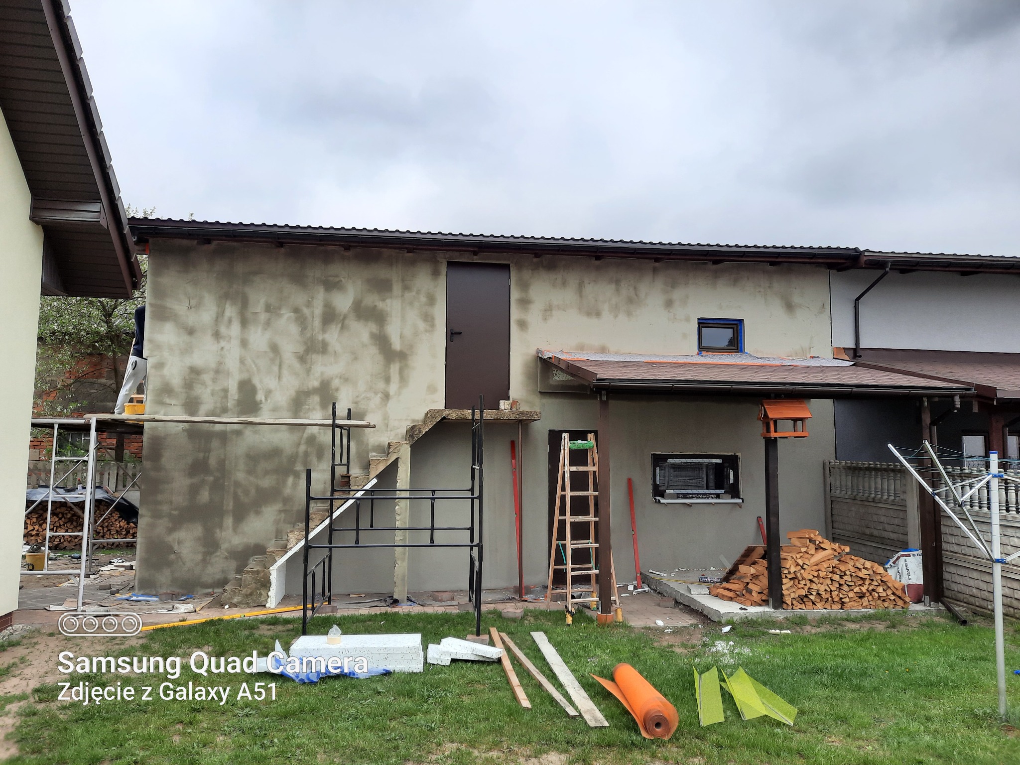 Budynek w trakcie prac elewacyjnych: surowy tynk na ścianie, rusztowanie, drabina oparta o ścianę, materiały budowlane na trawie, widoczne okno i drzwi wejściowe.
