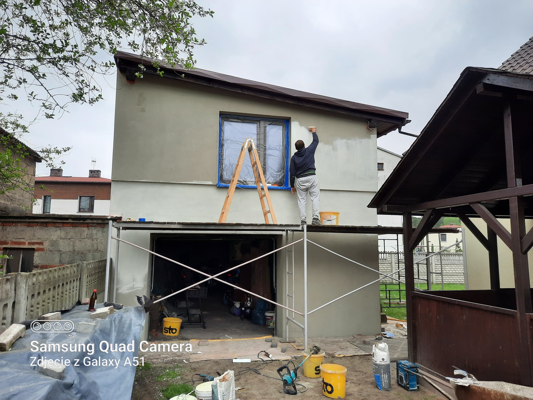 Mężczyzna maluje elewację domu z rusztowania, okno zabezpieczone folią, widoczne narzędzia i materiały budowlane.