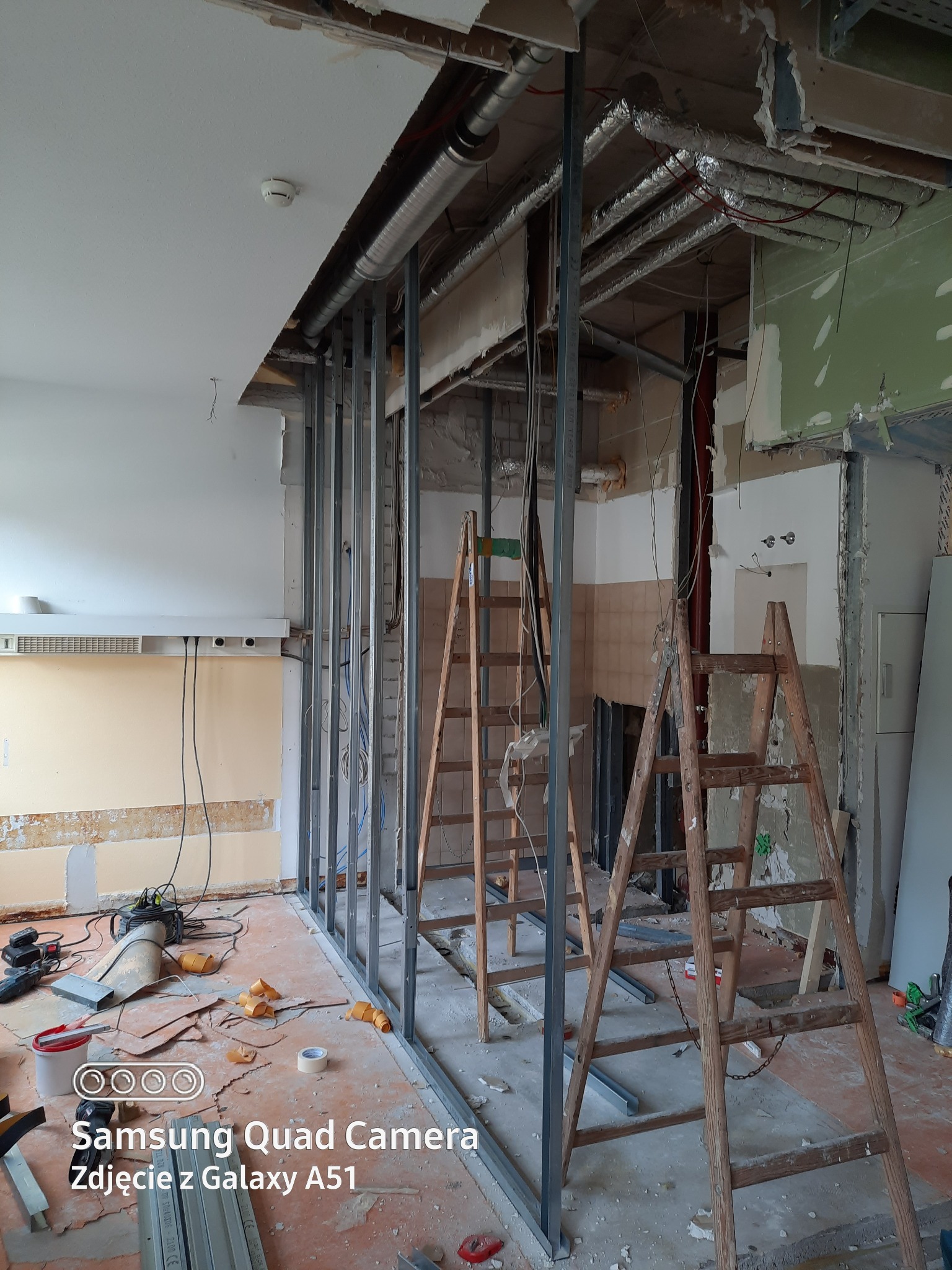 Wnętrze w trakcie remontu: stalowy szkielet ścianki działowej, dwie drewniane drabiny, widoczne instalacje pod sufitem, resztki materiałów budowlanych na podłodze.